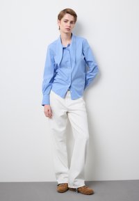 Persona apoyada contra una pared blanca, vistiendo una camisa azul con rayas y botones, pantalones blancos de pierna ancha y zapatos marrones con cuentas decorativas.