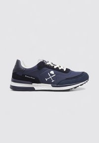 Zapatilla azul marino con cordones blancos, suela blanca, acentos negros y un logo de bates de polo cruzados y un casco en el lateral.