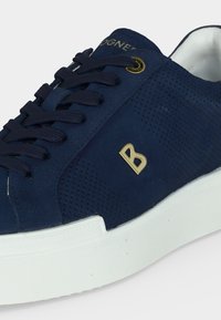 Baskets en daim bleu foncé avec côtés perforés, lacets noirs, logo lettre "B" doré, et semelle en caoutchouc blanche.