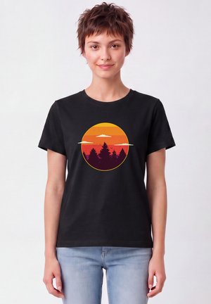Junge Person mit kurzen braunen Haaren trägt ein schwarzes T-Shirt mit einem runden Sonnenuntergang und Baumsilhouetten, kombiniert mit hellblauen Jeans.