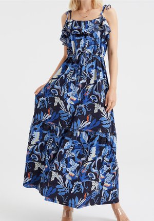 Femme portant une robe maxi sans manches à fleurs bleues avec un col à volants et des bretelles nouées sur les épaules, tenant l'ourlet de la robe.