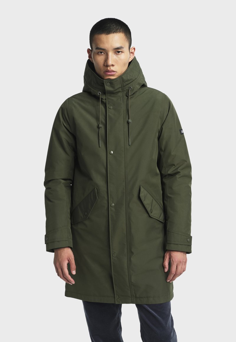 Modello maschile che indossa un lungo cappotto invernale verde oliva con colletto alto, lacci, bottoni a scatto e tasche a pattina, in piedi davanti a uno sfondo bianco.