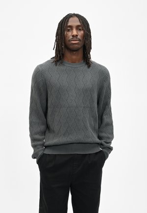 Mann mit schulterlangen Dreadlocks, der einen strukturierten grauen Pullover und schwarze Hose trägt, steht mit den Händen in den Taschen vor einem einfarbigen Hintergrund.