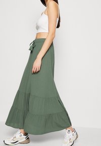 Jupe maxi verte, à volants, fabriquée en tissu léger avec une taille à cordon. Associée à un haut court blanc et des baskets de sport.