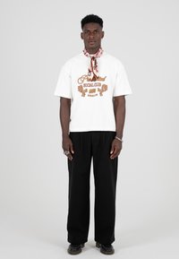 Hvit kortermet t-skjorte med brun grafisk tekst, parret med løse svarte bukser og et mønstret skjerf rundt halsen. Svarte sko.