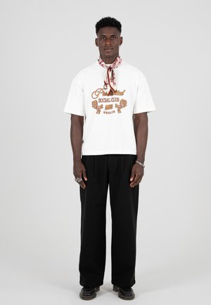 Camiseta blanca de manga corta con texto gráfico marrón, combinada con pantalones negros holgados y una bufanda estampada alrededor del cuello. Zapatos negros.