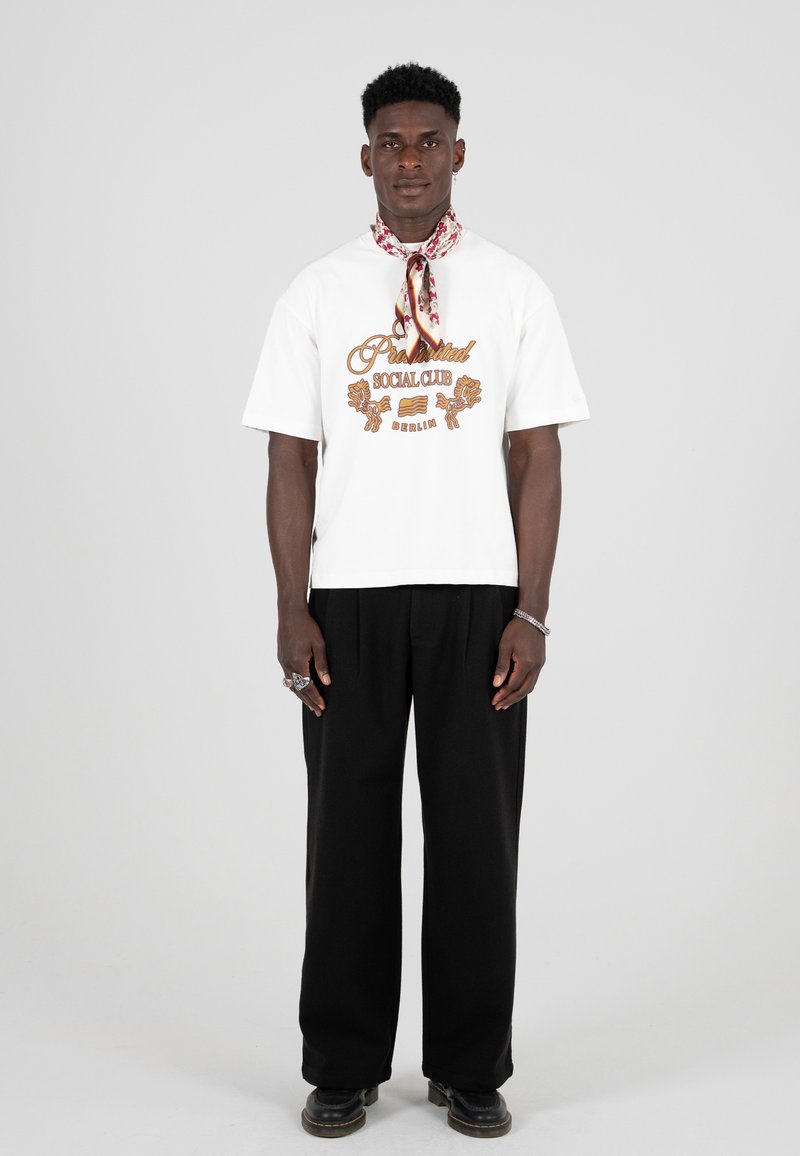 T-shirt bianca a maniche corte con testo grafico marrone, abbinata a pantaloni neri larghi e una sciarpa con motivo attorno al collo. Scarpe nere.