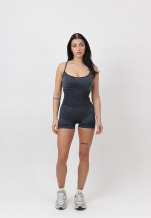 Kobieta z długimi ciemnymi włosami, ubrana w dopasowany, ciemnoszary, bezrękawnikowy kombinezon i białe tenisówki, stojąca na tle jednolitej białej ściany.