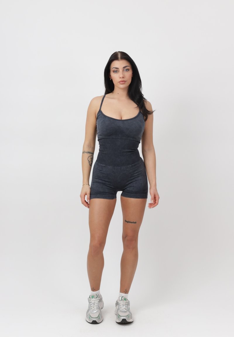 Mujer con cabello largo y oscuro que lleva un mono gris oscuro sin mangas ajustado y zapatillas blancas, de pie contra un fondo blanco liso.