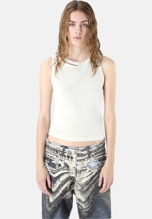 Junge Frau mit welligem Haar, die ein ärmelloses weißes Top und hoch taillierte schwarz-weiße gemusterte Jeans vor hellem Hintergrund trägt.