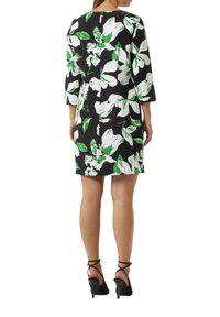 Blumenkleid mit schwarzem Hintergrund, großen weißen und grünen Blumen, Dreiviertelärmeln und knielangem Schnitt. Schwarze High Heels.