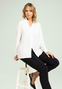 Witte blouse met lange mouwen en een kraag, gecombineerd met zwarte broek. Glad materiaal, ontspannen pasvorm, minimale details.