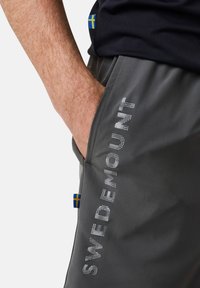 Grijze shorts met een gladde textuur, voorzien van de tekst "SWEDEMOUNT" in reflecterende stippen langs de zijkant en een accent van de Zweedse vlag.