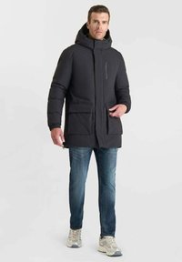 Parka nera isolata con una tasca sul petto con zip, due ampie tasche frontali, polsini elasticizzati e cappuccio, indossata sopra jeans blu e sneaker.
