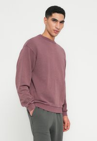 Lila Baumwoll-Sweatshirt mit Rundhalsausschnitt und gerippten Bündchen, auf der Brust mit einem dezenten Logo versehen. Kombiniert mit grauen Jogginghosen.