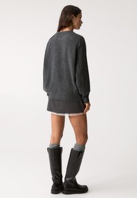 Temno siva oversize pulover s rebrastimi manšetami, v kombinaciji s sivo krilo in črnimi škornji do kolen. Model je prikazan s hrbta.