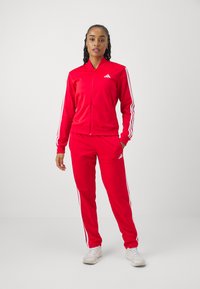 Červený teplákový komplet s bielymi pruhmi, sťahovacia bunda a zodpovedajúce nohavice. Vyrobené z hladkej tkaniny s logom Adidas na oboch kusoch.