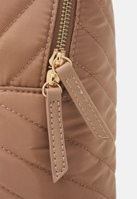 Två guldfärgade dragkedjehandtag med beige sydda läderlappar på en kviltad brun tygyta.