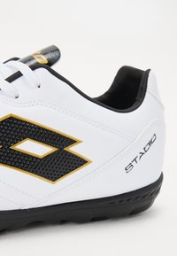 Baskets blanches avec un logo noir et or, un panneau noir texturé et des accents noirs sur le talon avec la marque "STADIO". Semelle en caoutchouc.