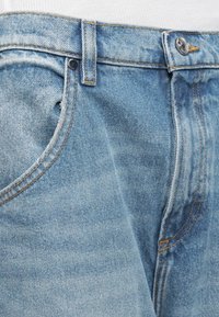 Φωτεινό γαλάζιο τζιν παντελόνι με ορατή μπροστινή τσέπη, θηλιά για ζώνη και ασημένιο κουμπί κλεισίματος σε λευκό υφασμάτινο φόντο.