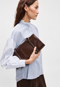 Καφέ δερμάτινο πορτοφόλι με λεία υφή, διακοσμητικό ρέλι και λεπτομέρεια με πόρπη, κρατημένο από ένα άτομο που φοράει ριγέ μπλούζα.