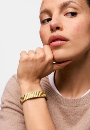 Donna che poggia il mento sul pugno, indossa un maglione beige e un bracciale dorato scanalato al polso, guarda di lato su uno sfondo semplice.