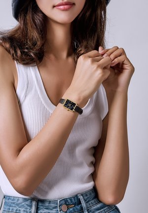 Reloj rectangular negro con detalles dorados, montado sobre una correa de cuero negra. El modelo lleva una camiseta sin mangas blanca de canalé y pantalones cortos de denim.