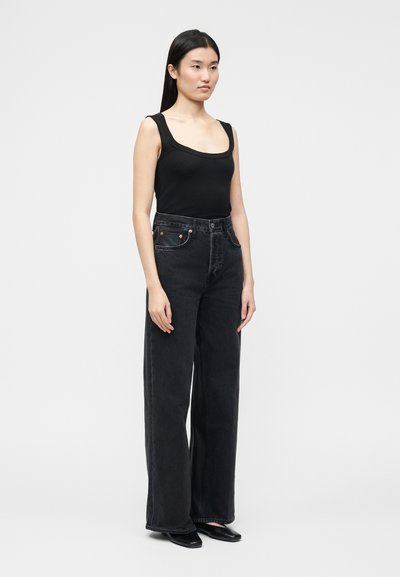 Γυναίκα με ίσια μαύρα μαλλιά, που φορά μαύρο τοπ χωρίς μανίκια, ψηλόμεσα φαρδιά μαύρα τζιν και μαύρα παπούτσια, στέκεται μπροστά σε απλό φόντο.