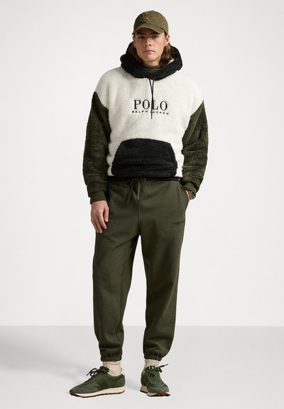 Haut à capuche blanc et olive, doublé en polaire, avec des accents noirs et un détail de logo, associé à un jogging olive et des baskets vertes. Ensemble vestimentaire décontracté.