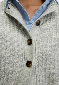 Person trägt ein hellblaues Hemd mit Knöpfen unter einer dicken, hellgrauen gestrickten Strickjacke mit großen braunen Knöpfen.