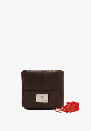 Square padded dark brown bag with white label and separate detachable bright red strap with hooks on white background.