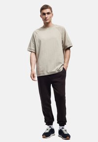 Beige oversized T-shirt og sorte sweatpants. T-shirten har en rund hals og korte raglanærmer. Sneakers er marineblå med hvide detaljer og snørebånd.