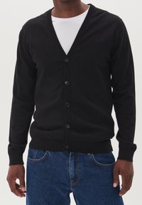 Černý cardigan s výstřihem do V, který má zapínání na knoflíky, žebrované manžety a lem. Nosený přes bílou košili, v kombinaci s modrými džíny.