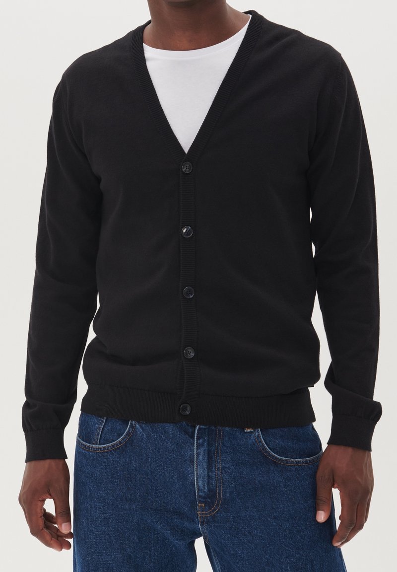 Černý cardigan s výstřihem do V, který má zapínání na knoflíky, žebrované manžety a lem. Nosený přes bílou košili, v kombinaci s modrými džíny.