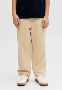 Enfant portant un pantalon large beige, des baskets blanches avec des semelles en gomme, et un haut bleu marine foncé, debout devant un fond clair uni.