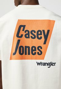 Camiseta de algodón blanca que presenta un gran rectángulo naranja con "Casey Jones" en texto negrita azul marino y el logotipo de "Wrangler" en negro en la esquina inferior derecha.