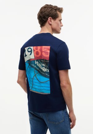 Mand, der vender mod højre iført navy T-shirt med stort farverigt grafisk patch på ryggen, der viser "1985" og tekst med rebdesign.