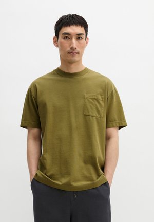 Man draagt een olijfgroen T-shirt met korte mouwen en ronde hals met borstzakje, handen in de zakken, tegen een lichte effen achtergrond.