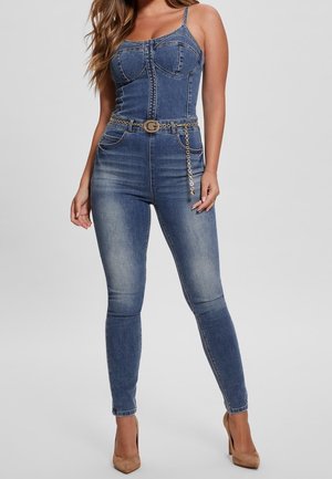 Kobieta ubrana w dopasowany niebieski kombinezon dżinsowy na cienkich ramiączkach, z ozdobnym łańcuchowym paskiem i brązowymi butami na wysokim obcasie na jednolitym tle.