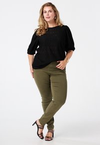 Zwarte blouse met textuur en puffed mouwen, gecombineerd met olijfgroene strakke broek en zwarte sandalen met banden. Eenvoudige accessoires maken de look compleet.
