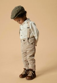 Beige corduroy broek met bretels, wit knoopshirt en bruine veterschoenen; een olijfgroene platkape maakt de outfit compleet.