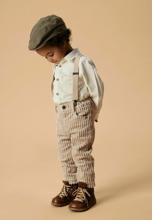 Beige fløjl bukser med seler, hvid skjorte med knapper og brune snørebåndsstøvler; en olivengrøn flad kasket fuldender outfittet.