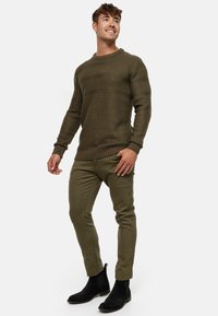 Olivgrüner Strickpullover mit strukturiertem Muster, gerippten Bündchen und Saum; kombiniert mit olivgrünen Slim-Fit-Hosen und schwarzen Stiefeletten.