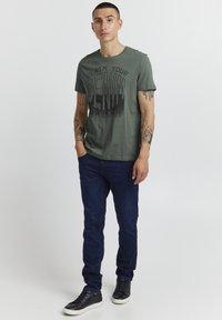 Grønn bomull t-skjorte med fet svart tekst "CLAIM YOUR DENIM". Båret med mørkeblå jeans og svarte joggesko. Tatoveringer synlige på armene.