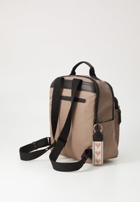 Zaino in tessuto beige con dettagli neri, doppio manico superiore, spallacci regolabili, tasca frontale con zip e etichetta decorativa sul lato.