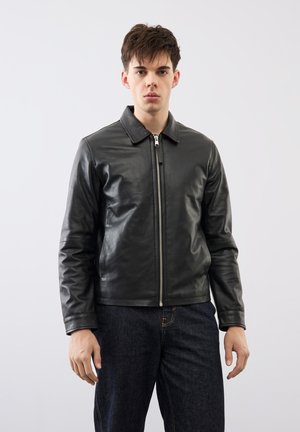 Ung mand iført en sort læderjakke med lynlås og mørke jeans, stående foran en ensfarvet lys baggrund og vendt fremad.