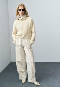 Maglione crema a coste con collo alto, abbinato a pantaloni beige larghi e scarpe bianche a punta, accessoriato con occhiali da sole neri.
