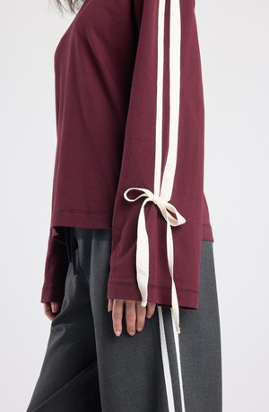 Bordeauxrotes Langarmoberteil mit weißen Seitenstreifen und gebundenen Akzenten am Handgelenk, kombiniert mit grauer Hose mit weißen Seitendetails.