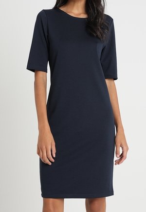 Femme portant une robe bleu marine longueur genou avec des manches aux coudes et un col rond, debout devant un fond uni.