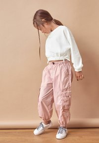 Biała, teksturowana bluza z regulowanym dołem, zestawiona z różowymi spodniami cargo z bocznymi kieszeniami i elastycznymi mankietami. Białe sneakersy z wzorzystymi sznurowadłami.